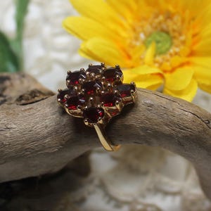 May include: A gold-toned ring featuring a cluster of deep red gemstones. The ring's design showcases a floral arrangement of the red stones, set against a natural wood background. A yellow flower is visible in the background.