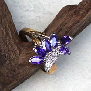 May include: A gold-toned ring featuring a floral design with marquise-cut purple gemstones and small white stones. The ring's band is a simple gold color, and the gemstones are arranged in a cluster.