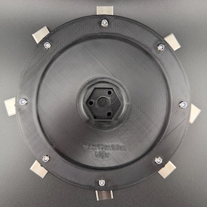 Peut inclure: Un composant circulaire noir imprimé en 3D doté d'une douille hexagonale centrale et de huit languettes rectangulaires métalliques régulièrement espacées, fixées par de petits boulons sur le pourtour. La surface présente une texture fine.