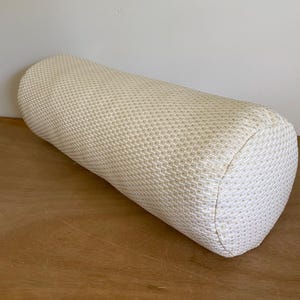 May include: A cylindrical bolster pillow with a textured, off-white fabric. The pillow is approximately 18 inches long and has rounded ends. The fabric has a subtle geometric pattern, and the pillow is resting on a wooden surface.