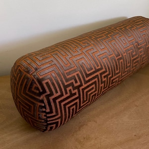 May include: A cylindrical bolster pillow with a geometric pattern in shades of brown and orange. The pattern is a repeating maze-like design. The pillow is made of a soft, textured fabric.