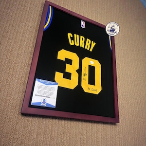 K&ouml;nnte beinhalten: Gerahmtes schwarzes Basketballtrikot mit dem Namen "CURRY" und der Nummer "30" in Gelb. Das Trikot ist signiert und enth&auml;lt "4x Champ". Der Rahmen ist dunkelburgunderfarben.