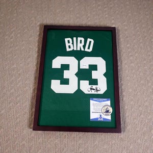 Könnte beinhalten: Gerahmtes grünes Basketballtrikot mit dem Namen "BIRD" und der Nummer "33" in Weiß. Das Trikot ist signiert und enthält ein Beckett-Echtheitszertifikat. Der Rahmen ist dunkelbraun.