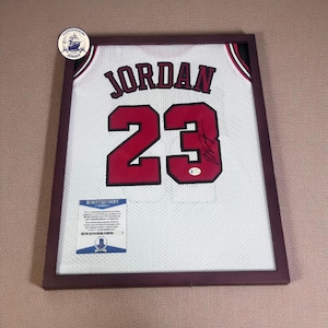 Può includere: Maglia da basket bianca incorniciata con il nome "JORDAN" e il numero "23" in rosso. La maglia è firmata e include un certificato di autenticità Beckett. La cornice è di colore marrone scuro.