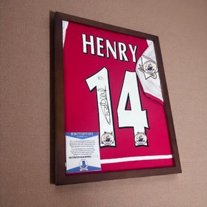 Può includere: Maglia rossa incorniciata con il nome "HENRY" e il numero "14" in bianco. La maglia è firmata e include toppe e un certificato di autenticità. La cornice è marrone scuro.