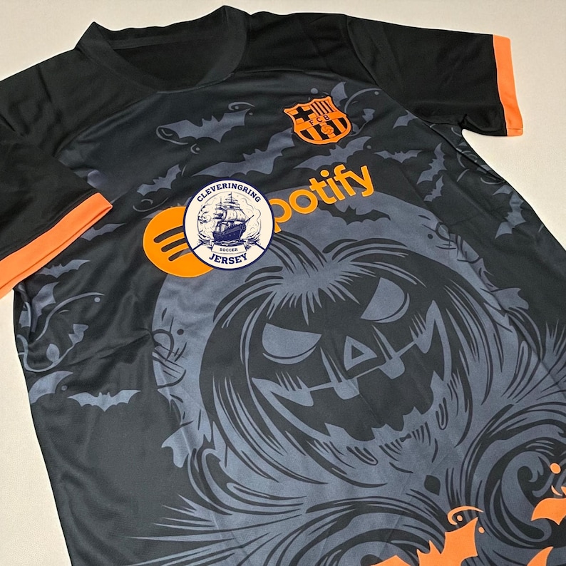 Op de afbeelding: Zwart voetbalshirt met oranje details, met een Halloween pompoen ontwerp en vleermuis silhouetten. Het shirt toont het Spotify logo en een team embleem. De mouwen hebben oranje manchetten.