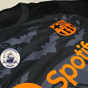 Op de afbeelding: Close-up van een zwart voetbalshirt met een oranje teamlogo en het woord "Spotify" in grote oranje letters. De trui heeft een subtiel grijs patroon en een "Cleveringring Soccer Jersey" logo.