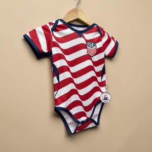 Pode incluir: Um body de bebé de manga curta com um padrão de riscas onduladas vermelhas e brancas e acabamentos em azul escuro. O peito apresenta um emblema em forma de escudo com o texto USA. Uma pequena etiqueta circular com um navio está anexada.