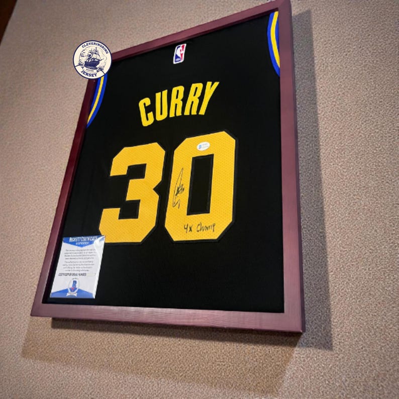 K&ouml;nnte beinhalten: Gerahmtes schwarzes Basketballtrikot mit dem Namen "CURRY" und der Nummer "30" in Gelb. Das Trikot ist signiert und enth&auml;lt den Text "4x Champ". Der Rahmen hat eine dunkelrotbraune Farbe.