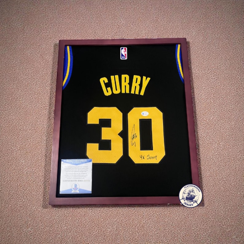 K&ouml;nnte beinhalten: Gerahmtes schwarzes Basketballtrikot mit goldenen Buchstaben und Zahlen. Das Trikot tr&auml;gt den Namen "CURRY" und die Nummer "30". Es ist signiert und enth&auml;lt eine kleine Echtheitskarte. Der Rahmen ist dunkelburgunderfarben.