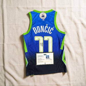 Puede incluir: Camiseta de baloncesto azul con el nombre "DONČIĆ" y el número "77" en blanco. La camiseta tiene ribetes verdes y un parche con un logotipo de barco. Un certificado está adjunto en la parte inferior.