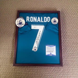 Może przedstawiać: Oprawiona turkusowa koszulka z nazwiskiem "RONALDO" i numerem "7" w kolorze białym. Koszulka jest podpisana i zawiera dwie okrągłe naklejki "Cleveringring Jersey". Rama jest ciemnobrązowa.
