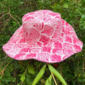 May include: A pink and white patterned sun hat with a seashell design. The hat has a wide brim and a rounded crown. The hat is made of fabric and is set against a background of green leaves and branches.