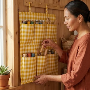 May include: A yellow and white gingham fabric wall organiser featuring six deep pockets. The organiser is suspended from a wooden rack and contains various spools of thread, small glass jars, and wooden tools stored within the individual pockets.