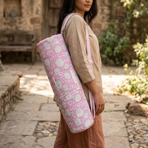 May include: A cylindrical yoga mat bag featuring a pink and white floral block print pattern. The bag has a rectangular front pocket and a long, adjustable shoulder strap made of matching pink and white striped fabric, designed for carrying.