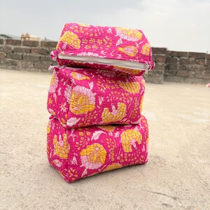 May include: Three stacked, rectangular cosmetic bags. Each bag is a vibrant pink with a repeating floral pattern in yellow and white. The bags have a zipper closure and are made of a quilted fabric.