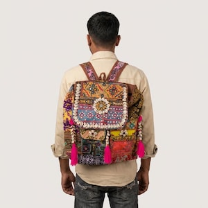 May include: A colorful patchwork backpack with a flap closure, adorned with intricate embroidery, seashells, and pink tassels. The backpack features a variety of patterns and colors, including yellow, blue, and red, and brown leather straps.