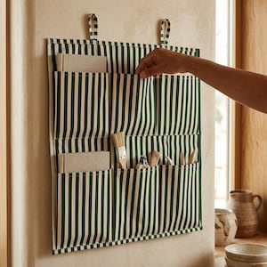May include: A wall-hanging fabric organiser featuring a vertical dark green and cream striped pattern. The organiser has six individual pockets, with two beige notebooks, a paintbrush, and wooden sculpting tools tucked inside the compartments.