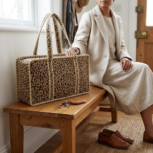 May include: A rectangular tote bag featuring a brown and black leopard print pattern with structured piping along the edges and two tall handles. A small brown leather coin pouch and a set of metal keys rest on the wooden surface nearby.