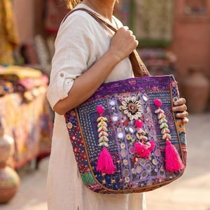 May include: A colorful tote bag with a brown leather strap. The bag features a vibrant design with purple, blue, and pink accents, including tassels, mirrors, and shell embellishments. The bag is held by a person.