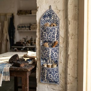 May include: A blue and white fabric wall organiser with three pockets, featuring a floral design. The pockets contain spools of thread, twine, and a wooden tool. The organiser is mounted on a textured, off-white wall.