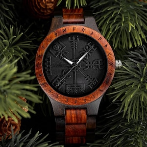 May include: A wooden wristwatch with a dark face featuring a compass design and runic symbols. The watch has a dark brown wooden case and band, with a lighter brown wooden bezel. The watch is surrounded by green pine needles and pine cones.