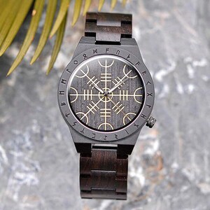 Puede incluir: Un reloj de pulsera de madera negra con una esfera negra y detalles dorados. La esfera presenta un diseño circular con runas alrededor del borde y un diseño de brújula en el centro.