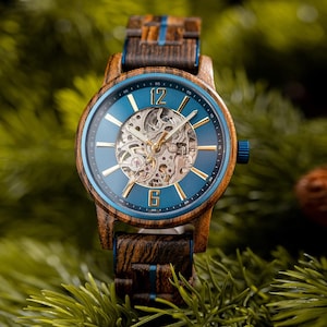 Puede incluir: Un reloj de pulsera de madera con esfera azul y movimiento mecánico visible. El reloj tiene agujas e indicadores dorados, y una correa de madera con detalles azules. La caja del reloj mide aproximadamente 4,4 cm de diámetro.