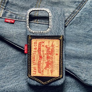 Op de afbeelding: Een denim telefoonhoesje met een gestikte rand rond de camera-opening. De hoes heeft een Levi Strauss & Co.-label met rode en bruine accenten. De achtergrond is een denim stof.