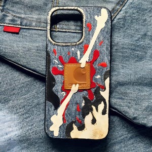 May include: A denim-textured phone case with a splatter design in red, white, and black. The case features a brown leather patch with the Carhartt logo. The case is designed to fit a smartphone.