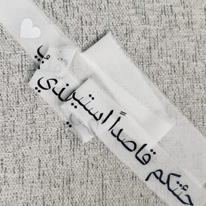 Peut inclure: Tissu blanc avec texte arabe noir et un petit cœur blanc. Le texte se lit « Je viens vers toi ». Le tissu est superposé sur une surface grise texturée.