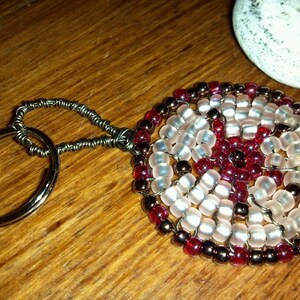 May include: A beaded keychain with a circular design featuring a swirl pattern of white and red beads. The keychain is attached to a silver key ring.