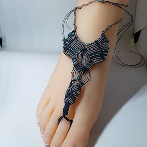 May include: A blue and gray macrame foot jewelry with green beads. The foot jewelry is tied around the ankle and has a decorative design that wraps around the toes.