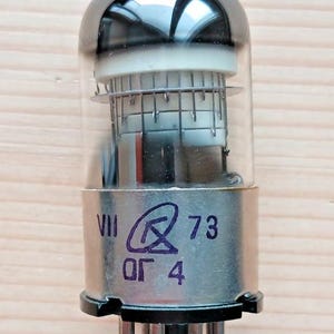 Puede incluir: Una válvula de vacío vintage con una bombilla de vidrio transparente y una base de metal plateado. El tubo tiene componentes internos visibles a través del vidrio y marcas que incluyen "VII", un símbolo, "73" y "OF 4".