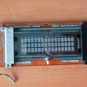 Pode incluir: Um componente eletrônico com uma placa de circuito retangular laranja. Possui uma grade de pequenos quadrados e pinos ao longo das bordas. Os fios se estendem de um lado, e um suporte de metal protege uma área de exibição central.