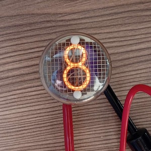 Puede incluir: Primer plano de un tubo Nixie que muestra el número "8" en naranja. El tubo está encerrado en una bombilla de vidrio transparente con una rejilla metálica y un punto blanco en la parte superior.