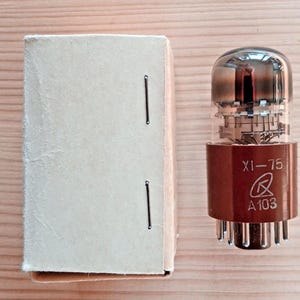 May include: A vintage vacuum tube with a brown base and clear glass top, labeled "XI-75" and "A103." Beside it is a beige cardboard box. The tube has multiple internal components and metal pins at the bottom. The items are on a light wood surface.