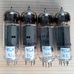 Op de afbeelding: Vier vintage vacuümbuizen met glazen behuizingen en metalen bases. Elke buis heeft een label met handgeschreven tekst, inclusief nummers en metingen zoals "45mA" en "11,1s". De buizen zijn verticaal gerangschikt op een licht houten oppervlak.