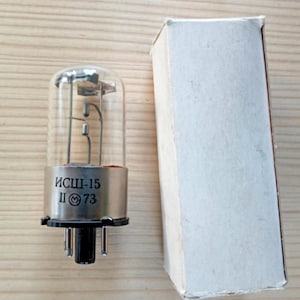 May include: A vintage vacuum tube with a clear glass bulb and metallic base, next to a plain white cardboard box. The tube has internal components visible through the glass. The base has text and numbers printed on it.