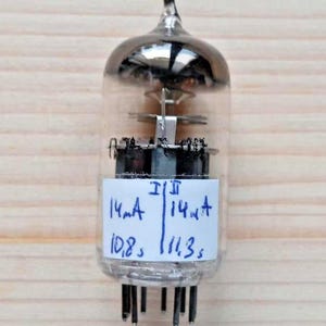 May include: A clear glass valve with a silver top and black base. A white label displays handwritten measurements: "14mA, 10.8s" and "14mA, 11.3s". The valve has multiple pins at the bottom and is set against a light wood background.