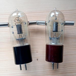 Puede incluir: Dos válvulas de vacío vintage con bombillas de vidrio transparente, conectores metálicos y bases cilíndricas. Una base es negra, la otra marrón. Los tubos tienen componentes internos visibles a través del vidrio.