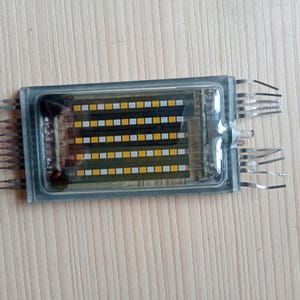 Puede incluir: Componente electrónico rectangular con una carcasa de plástico transparente. La estructura interna presenta filas de cuadrados amarillos, blancos y negros. Los pines metálicos se extienden desde los lados.