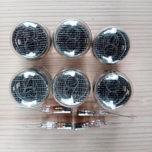 May include: Six vintage Nixie tubes with clear glass bulbs and wire mesh interiors. Below the tubes are six smaller glass components with metal pins and wires, likely the tube's connectors. The components are arranged on a light-colored wooden surface.