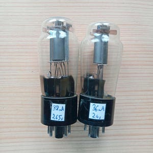 May include: Two vintage vacuum tubes with clear glass bulbs and black bases. Each tube has internal components and a label with handwritten numbers and letters. The tubes are on a light brown wooden surface.