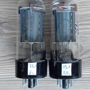May include: Two vintage vacuum tubes with clear glass bulbs and black bases. Each tube has a label with handwritten text, including "78mA" and "6 s" or "5.85 s". These are likely electronic components.