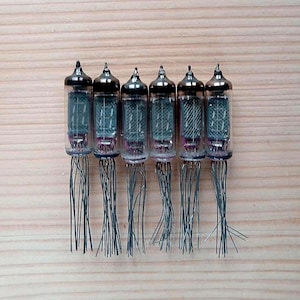 Puede incluir: Seis tubos de vacío vintage con carcasas de vidrio, tapas de metal y numerosos cables. Los tubos tienen un interior oscuro con componentes internos visibles. Están dispuestos en fila sobre una superficie de madera clara.