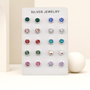 May include: A display of silver jewelry earrings with colourful gemstones. The earrings are arranged on a white card with the text "SILVER JEWELRY" at the top. The earrings have various colours, including green, blue, pink, and clear.