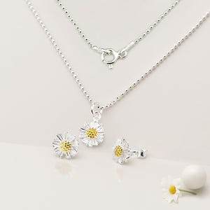 以下が含まれることがあります： シルバーのデイジーフラワーのジュエリーセット。ペンダント付きのネックレスと、お揃いのスタッドピアスが含まれます。 デイジーのペンダントとイヤリングは、白い花びらと黄色の中心部が特徴です。 ネックレスチェーンは繊細なボールチェーンです。
