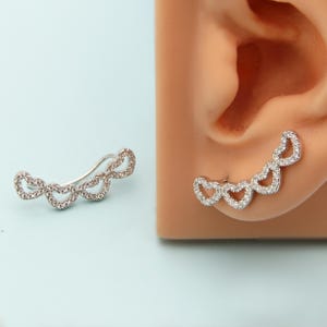 May include: A silver ear cuff featuring a series of heart-shaped designs, each encrusted with small, clear gemstones. The cuff curves along the ear, creating a delicate and sparkling accessory. The design is shown on a light blue background.