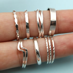 Może przedstawiać: Kolekcja srebrnych pierścionków o różnych wzorach. Pierścionki obejmują młotkowaną obrączkę, skręconą obrączkę, gładką obrączkę i obrączkę z falistą krawędzią. Pierścionki są prezentowane na palcu na jasnoniebieskim tle.